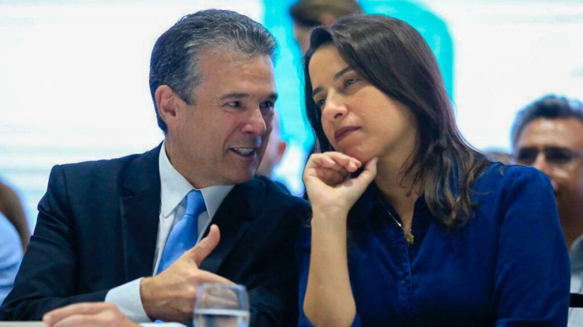 André de Paula fala sorrindo enquanto olha para Raquel Lyra, que escuta com uma mão no queixo, ambos estão sentados