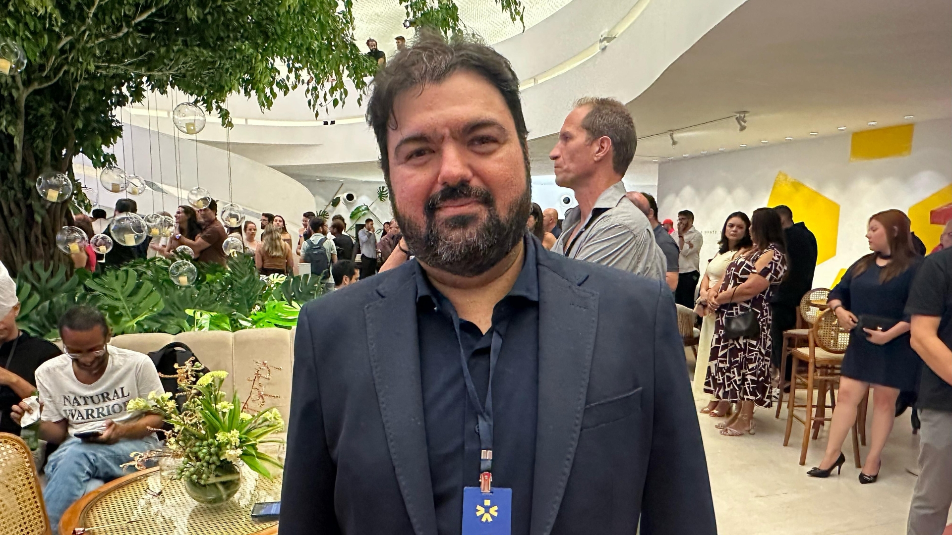 Um homem branco de cabelo curto castanho de barba e bigode usa um paletó com camisa social preta, ao fundo diversas pessoas