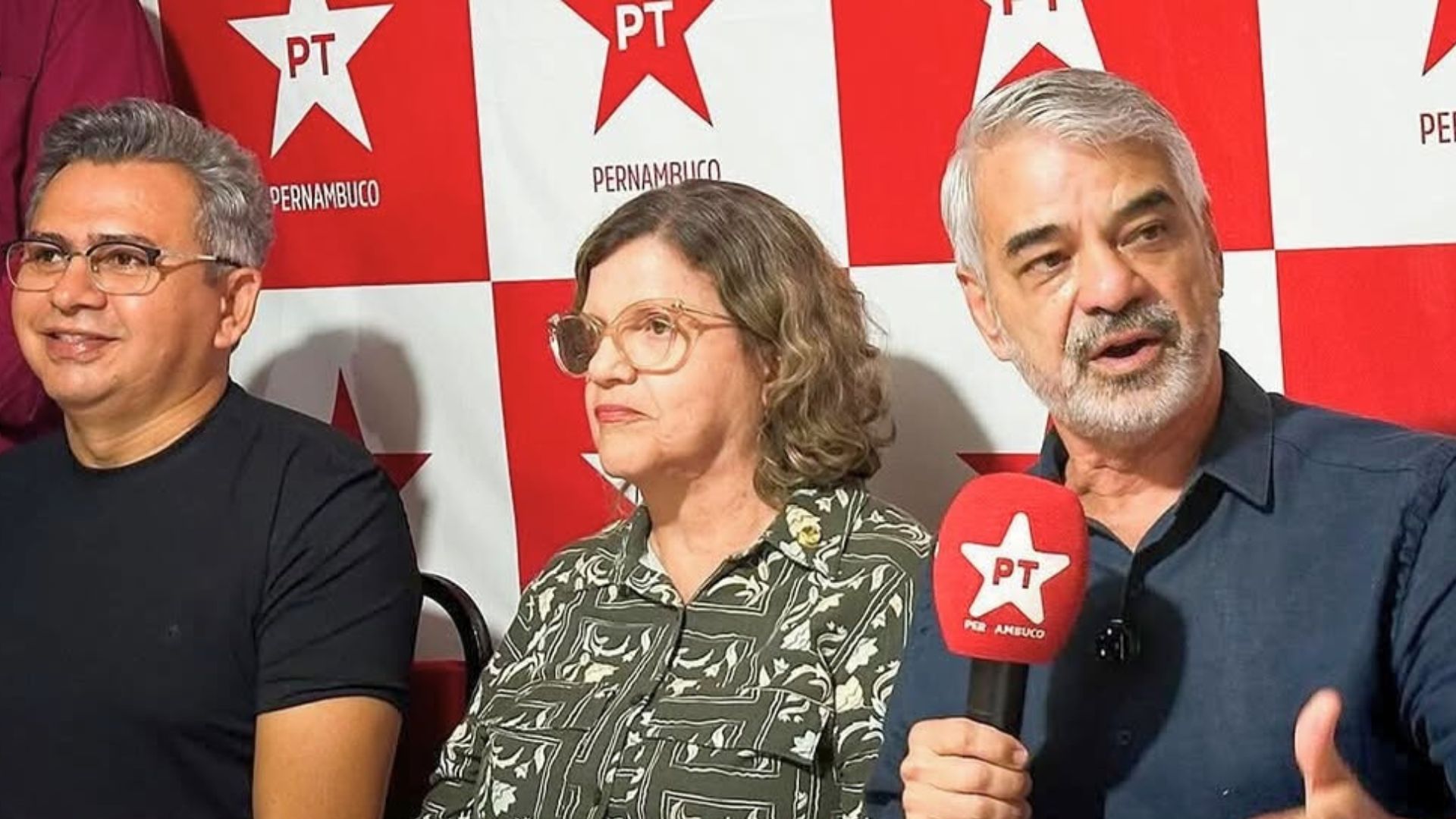Humberto Costa está sentado ao lado de Teresa Leitão, ele fala com um microfone com espuma vermelha e estrela escrito PT, ao fundo há a estampa do PT Pernambuco em vermelho e branco