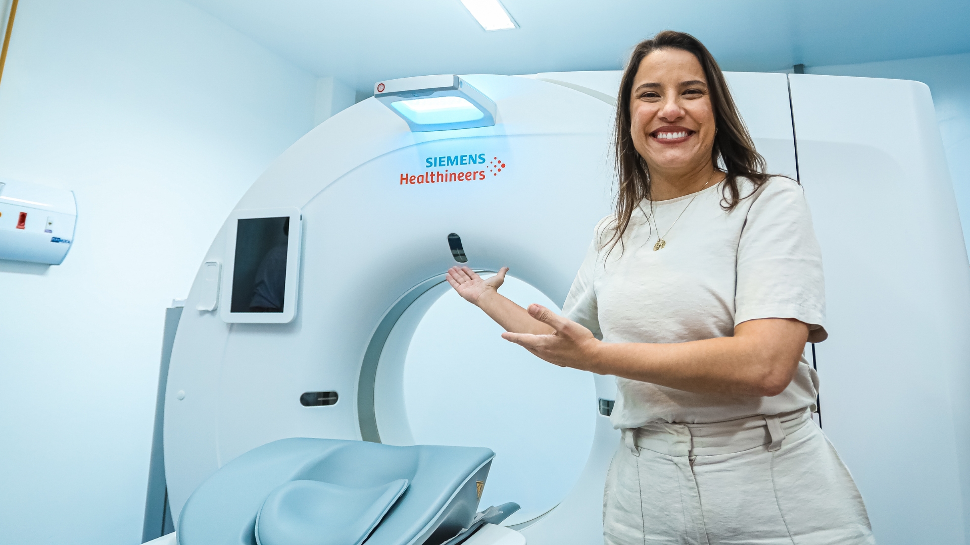 Raquel Lyra sorri e aponta para máquina de ressonância ao seu lado