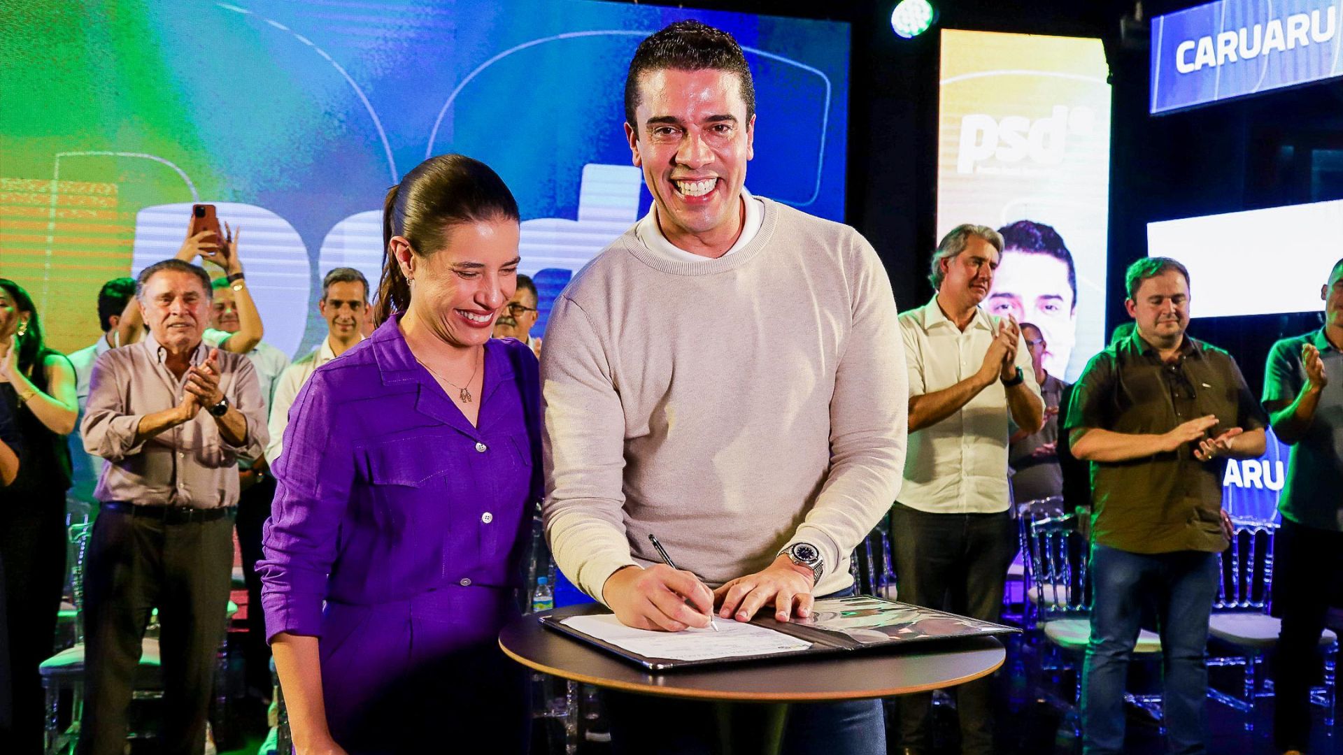 Rodrigo Pinheiro sorri enquanto assina documento, Raquel Lyra sorri e olha para documento. Ao fundo, pessoas batem palma, um deles é João Lyra