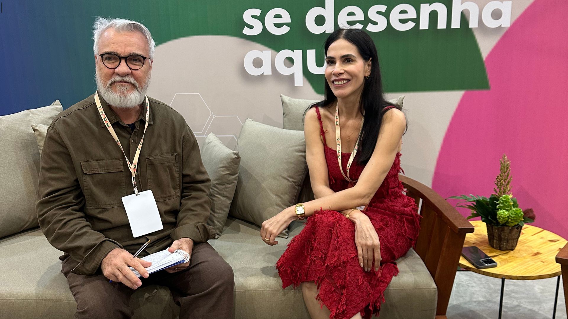 Jamildo Melo e Simone Nunes lado a lado sentados em um sofá, jamildo tem um caderno e caneta nas mãos