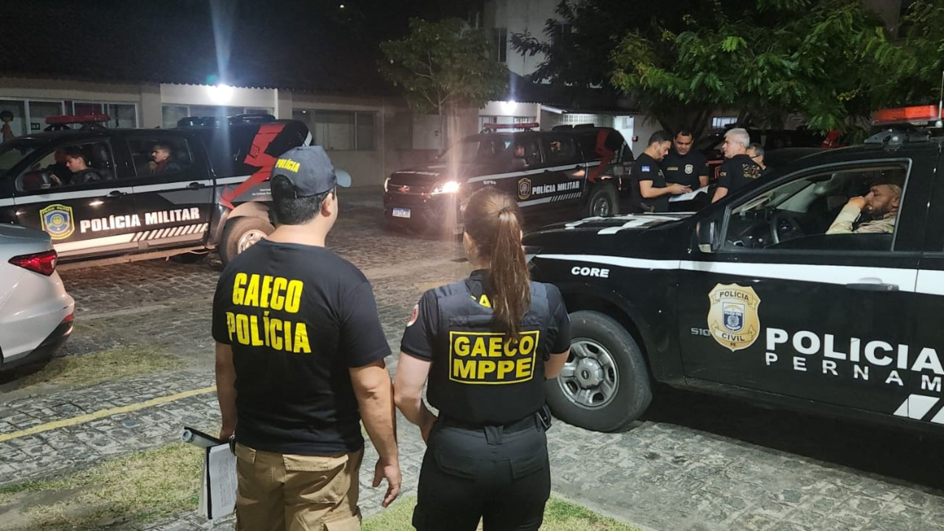 Um homem e uma mulher em pé de costas. Farda do homem está escrito "Gaeco polícia", da mulher está escrito "Gaeco MPPE". Ao fundo viatura da polícia militar. Está escuro