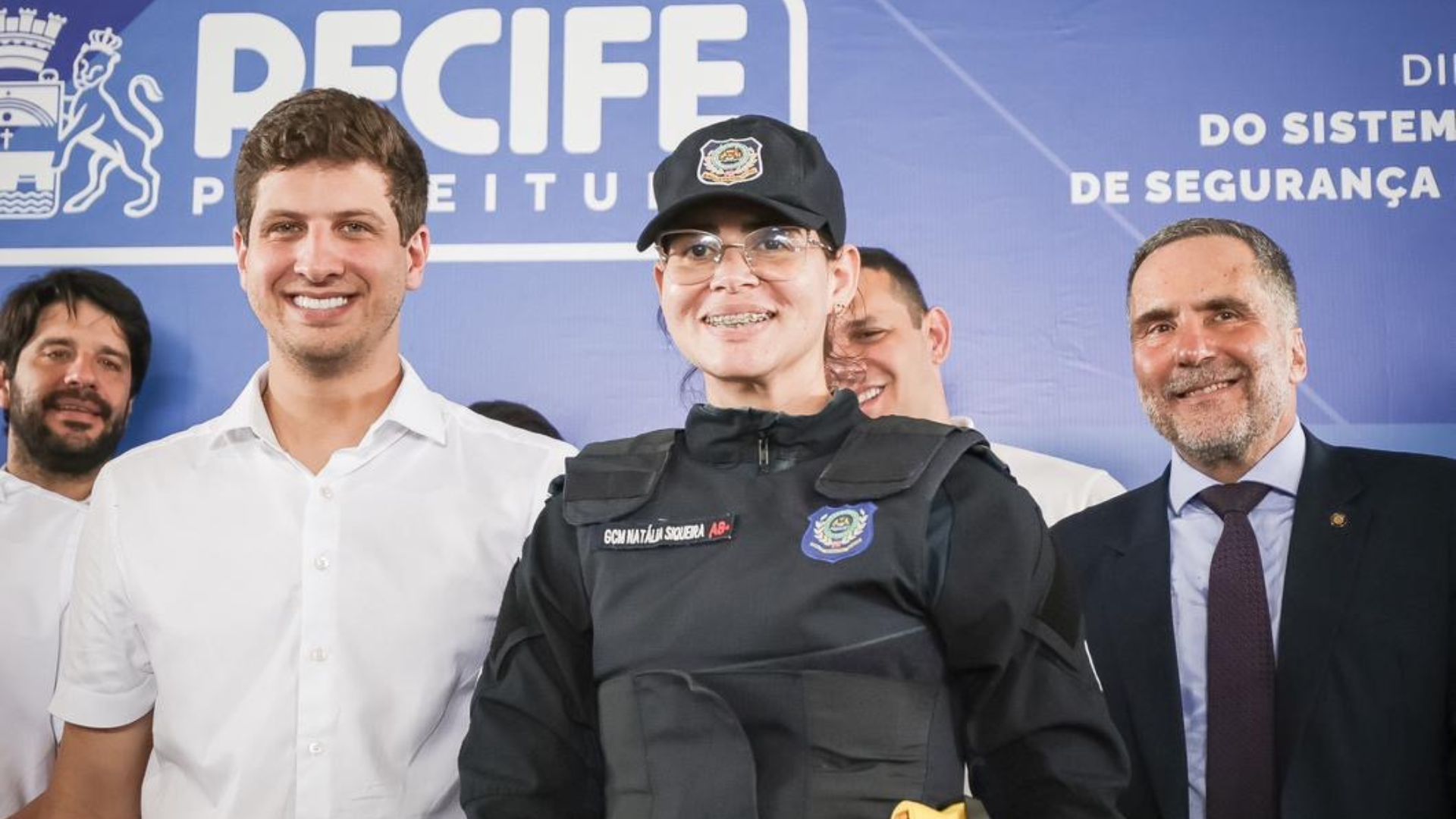 João Campos ao lado de uma mulher com farda estilo policial e boné da guarda municipal, ao fundo logo da prefeitura do recife e escrito ao lado sistema de segurança