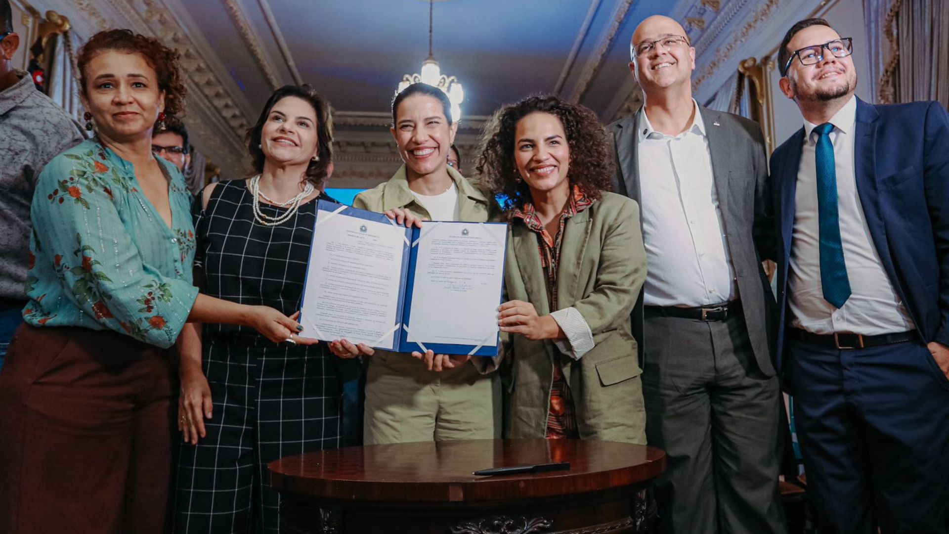 Raquel Lyra, Priscila Krause, Rosa Amorim seguram documento enquanto sorriem em pé, ao lado estão dois homens e uma mulher negra