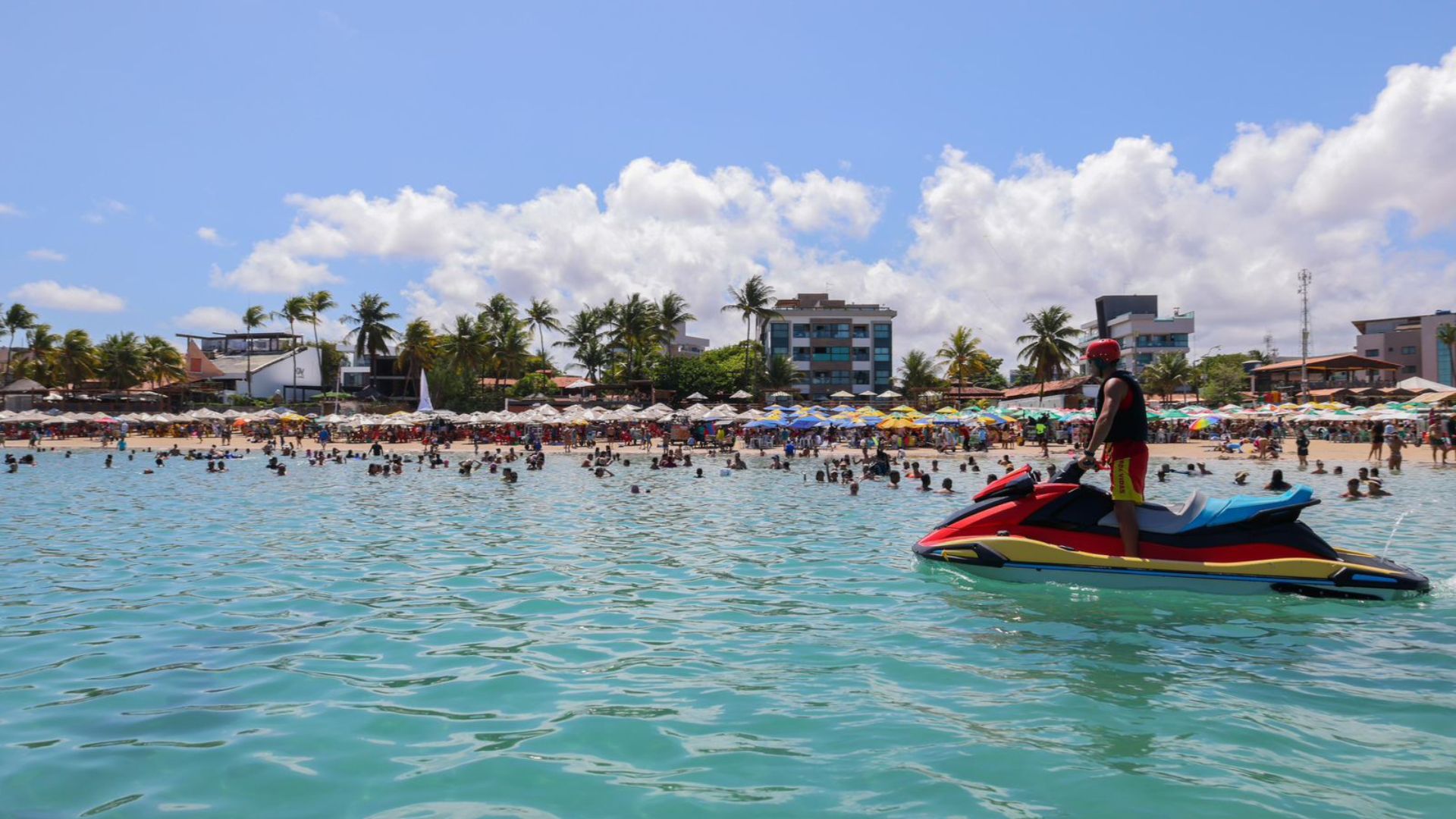 Mar azul com homem em jet ski, ao fundo diversas pessoas e praia