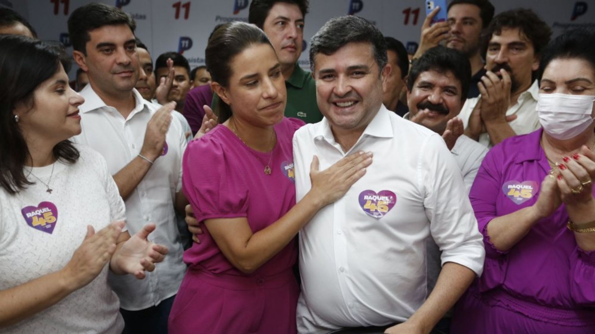 Raquel Lyra abraça Eduardo da Fonte, que sorri. Ele usa um adesivo de campanha de Raquel. Várias pessoas batem palma ao redor
