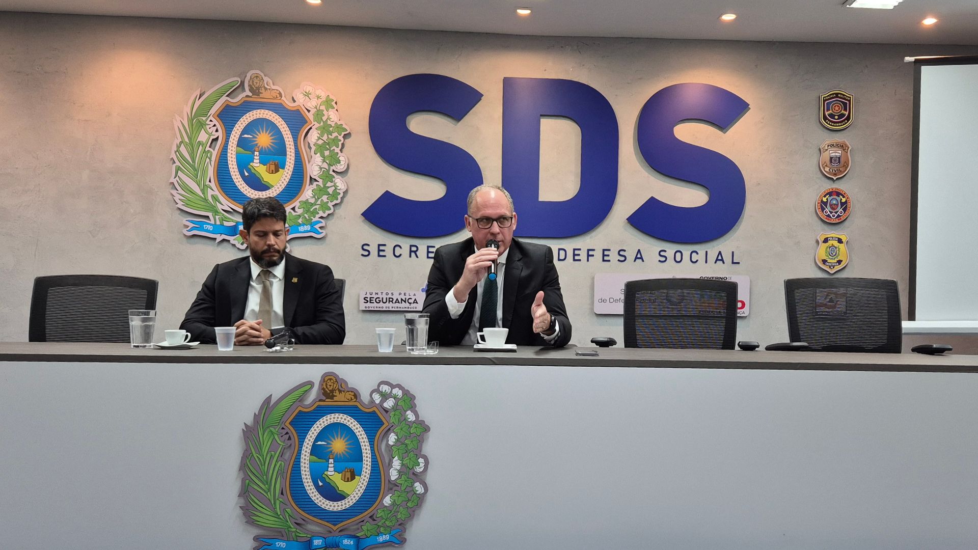 Alessandro e Felipe sentados em mesa com logo do governo de pernambuco, ao fundo simbolo da sds