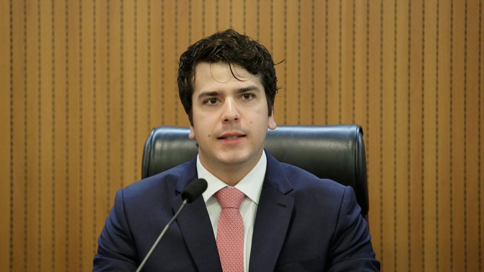 Antonio Coelho fala em microfone sentado em cadeira. Ele usa terno