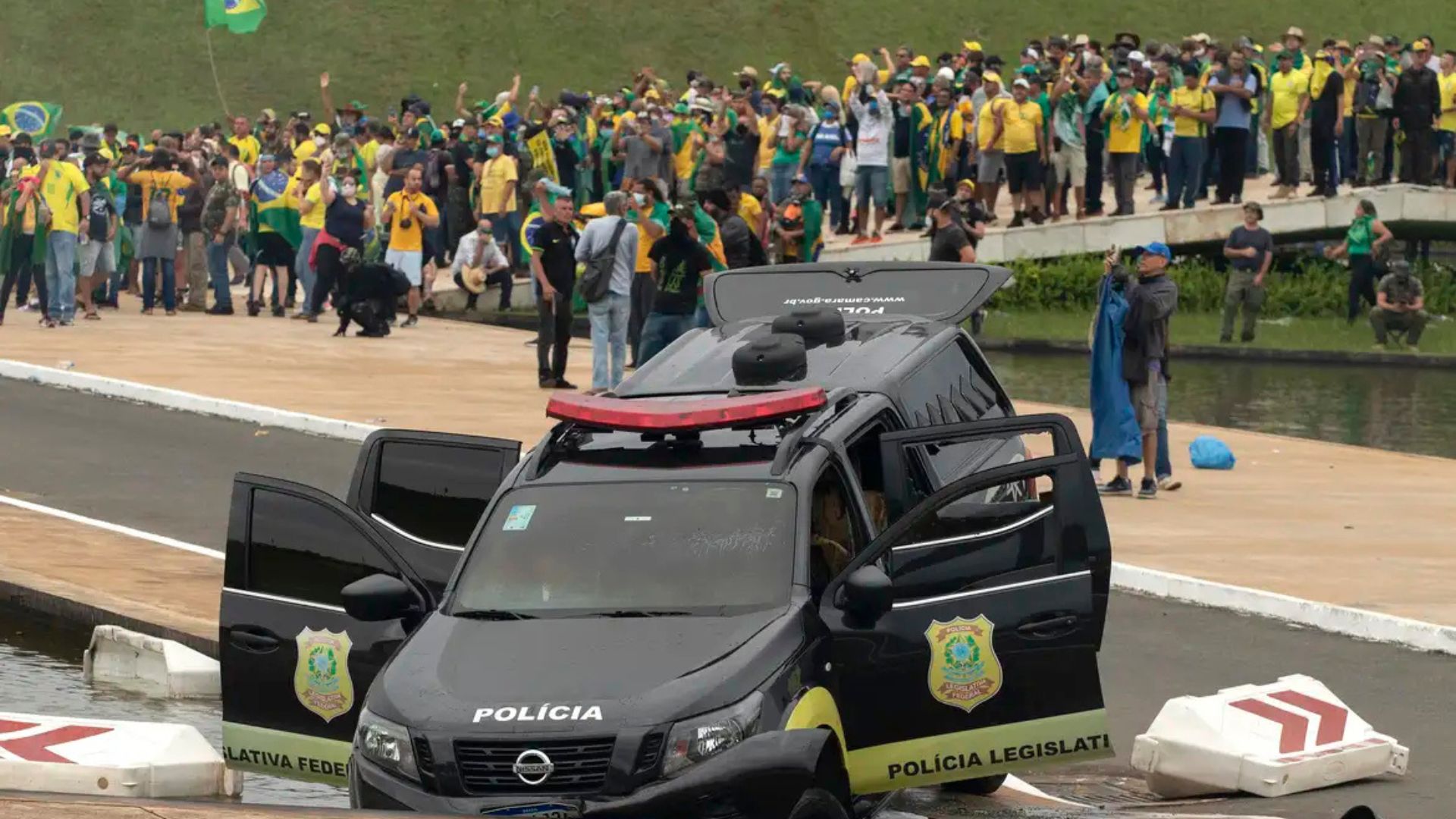 Viatura preta da polícia legislativa jogada em canal, malas e portas abertas. Ao fundo, manifestantes com camisas do Brasil, na Praça dos Três Poderes.