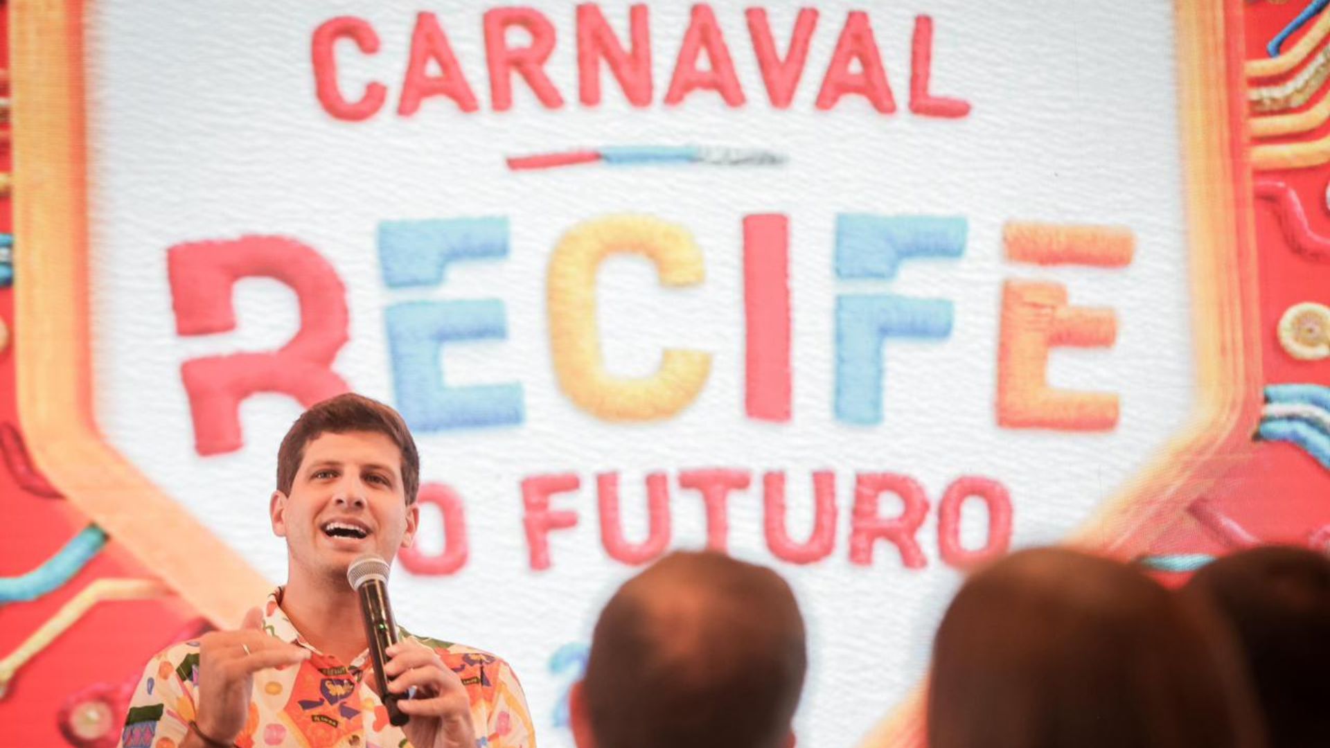 João Campos com camiseta florida fala em microfone. Ao fundo desenho escrito "Carnaval Recife do Futuro"