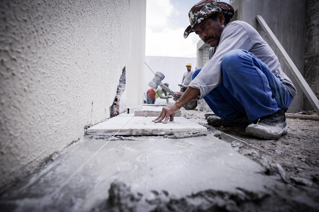 Homem negro, de calça jeans e camisa manga longa, martelando um prego em uma tábua.
