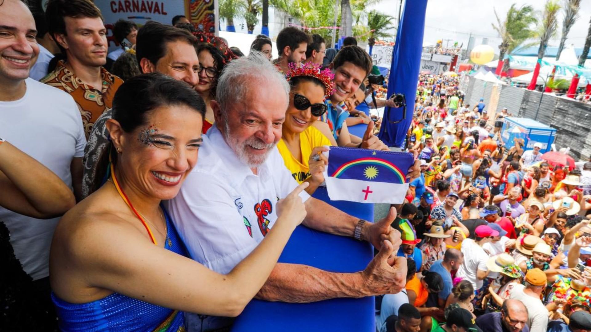 Raquel Lyra aponta para bandeira de Pernambuco pequena que Janja segura. Lula e João Campos sorriem. Ao fundo multidão embaixo. Todos usam roupas carnavalescas