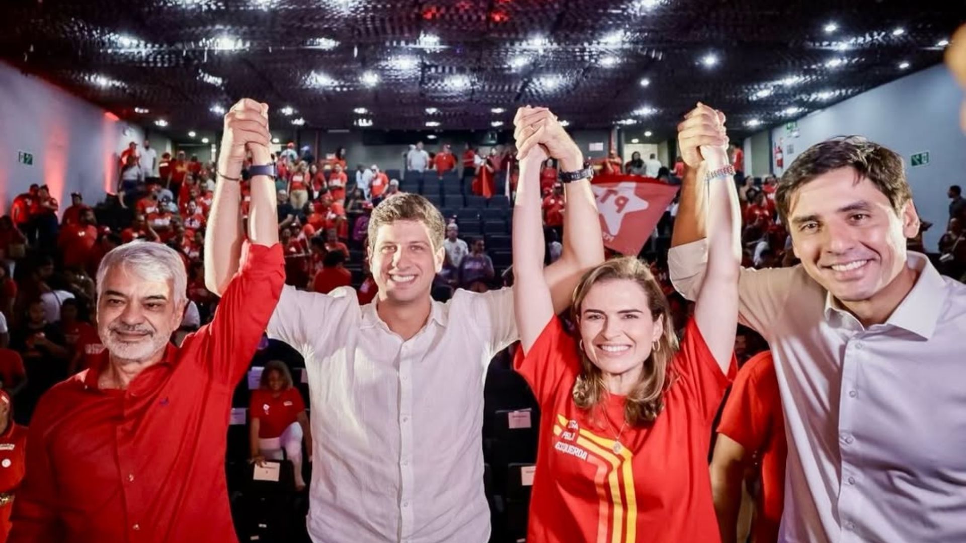 Humberto Costa, João Campos, Marília Arraes e Carlos Costa sorriem com as mãos erguidas unidas. Ao fundo diversas pessoas com roupas vermelhas e bandeira do PT