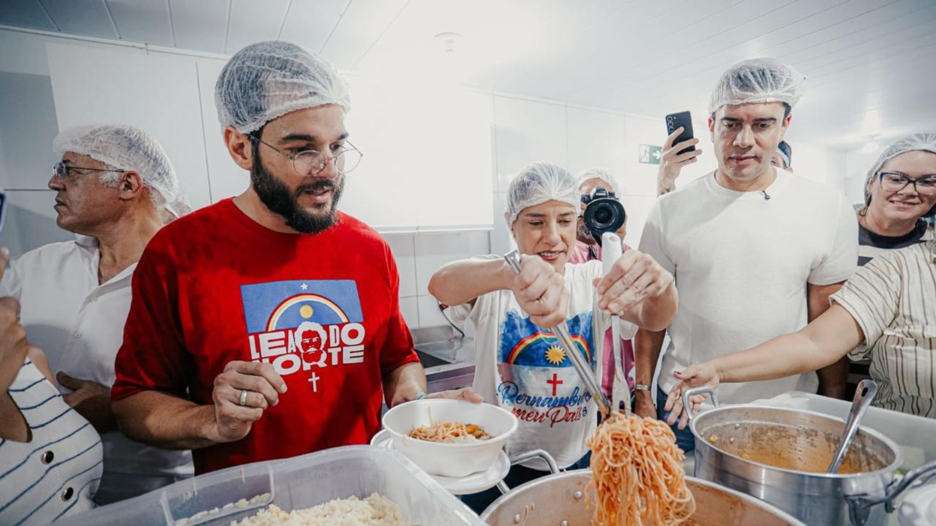 Todos de touca. Raquel Lyra serve um macarrão de panela. Túlio Gadelha, Rodrigo Pinheiro e outros olham para ação. Cenário de cozinha com diversas panelas