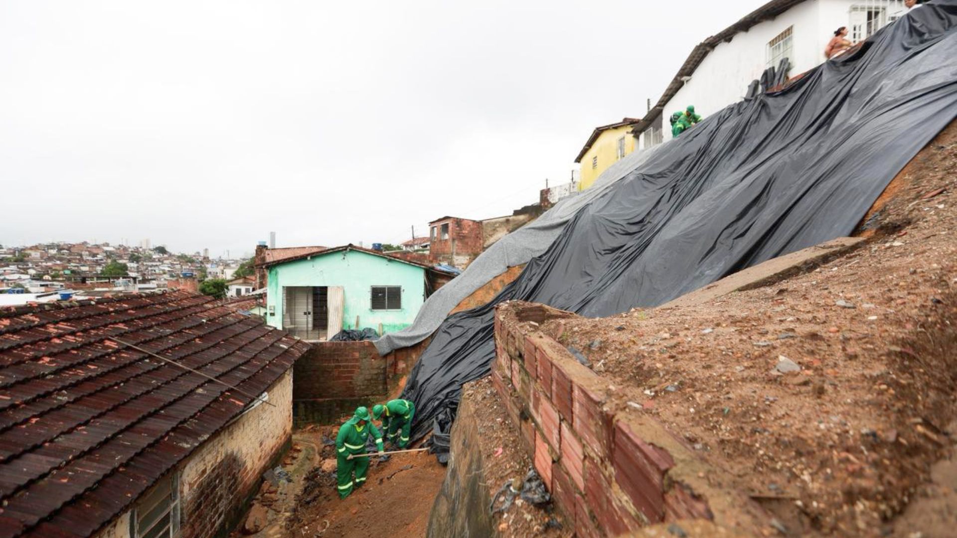 Encosta coberta por lona preta. Em cima e abaixo da encosta há casas