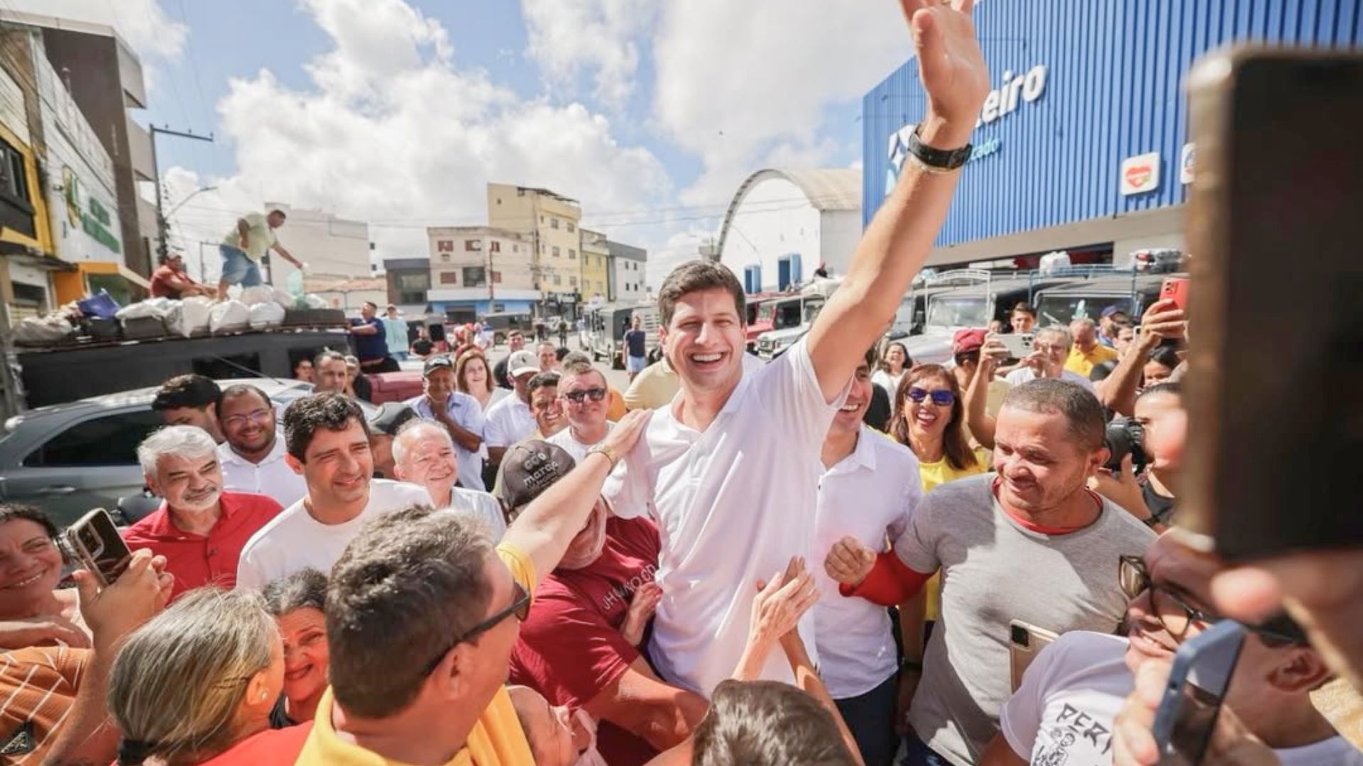 João Campos é levantado por um homem enquanto acena, multidão o segue. Entre pessoas estão Rodrigo Farias e Humberto Costa