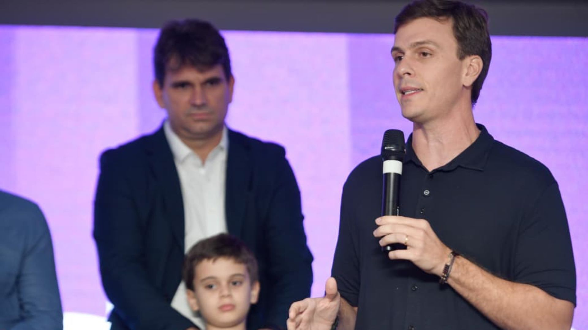 Miguel Coelho fala em Microfone. Ao fundo Marcelo Gouveia observa enquanto segura uma criança