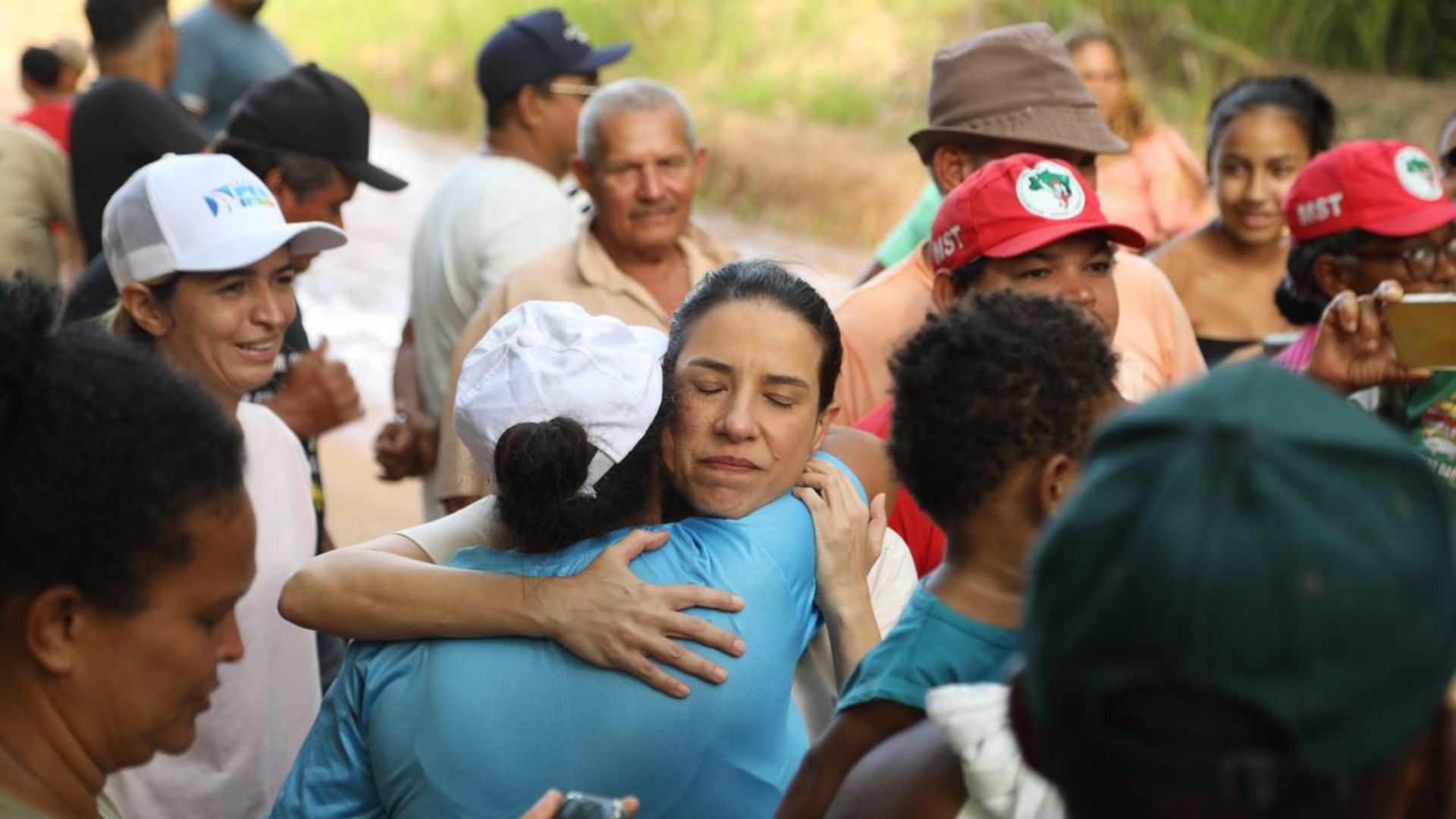 Raquel Lyra abraça mulher, governadora está de olhos fechados. Diversas pessoas de chapéus, algumas do MST estão ao redor. Ao fundo estrada de barro