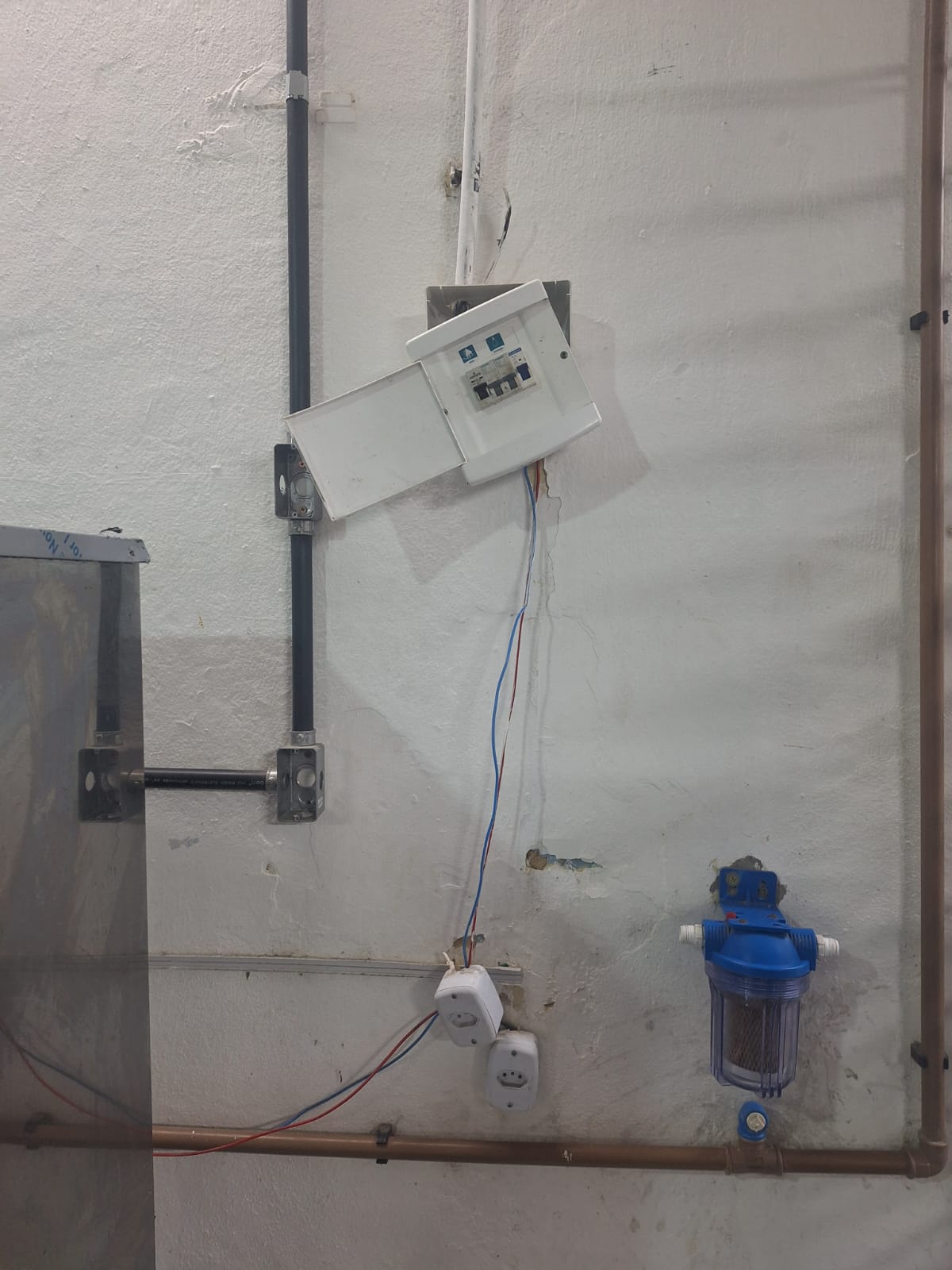 Quadro de disjuntores elétricos quebrado e pendurado na parede com fiação exposta acima de tomadas desalinhadas e um filtro de água.