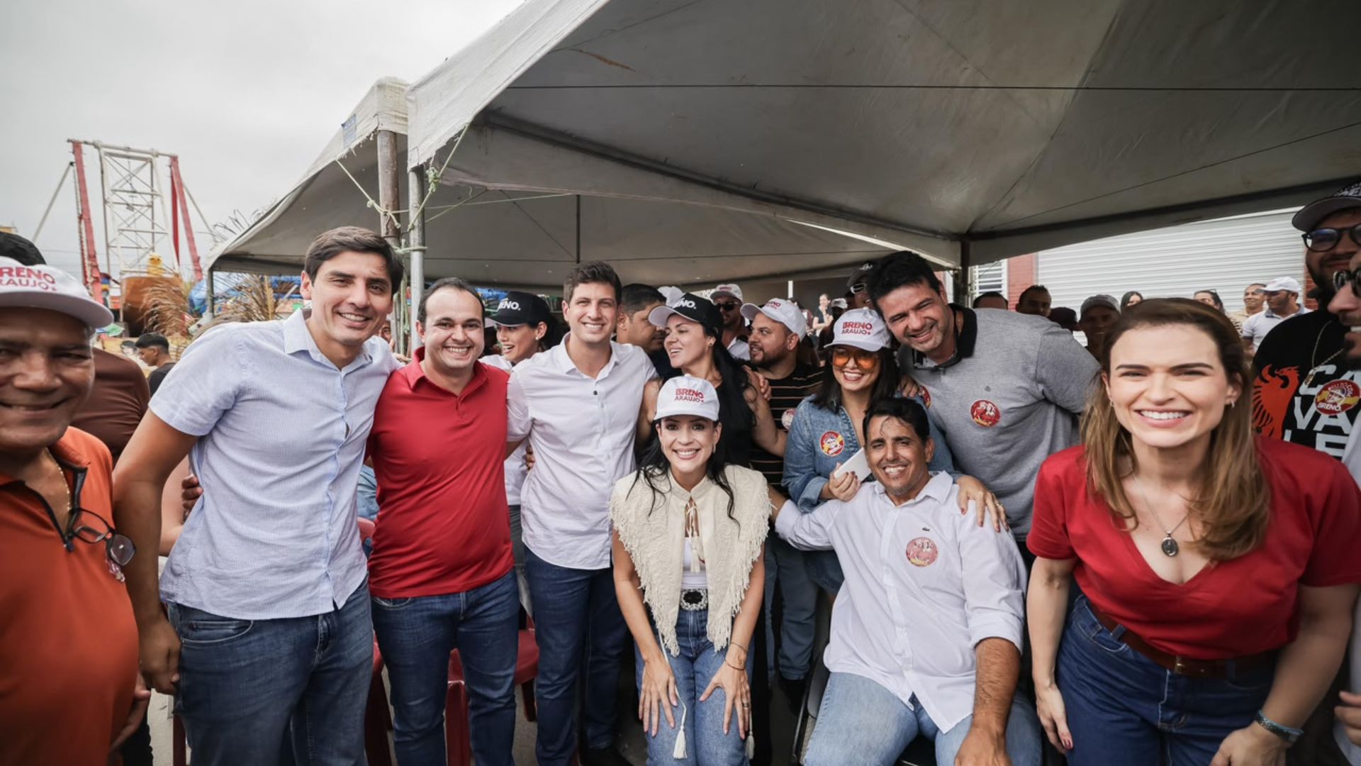 Grupo de pessoas, entre elas João Campos, Marília Arraes, Carlos Costa e Márcia Conrado sorrindo e posando para foto em evento ao ar livre sob tenda branca. Homens e mulheres vestindo camisas em tons de azul, vermelho e branco, alguns usando bonés com o nome de Breno Araujo. Ao fundo, público e estruturas de evento.