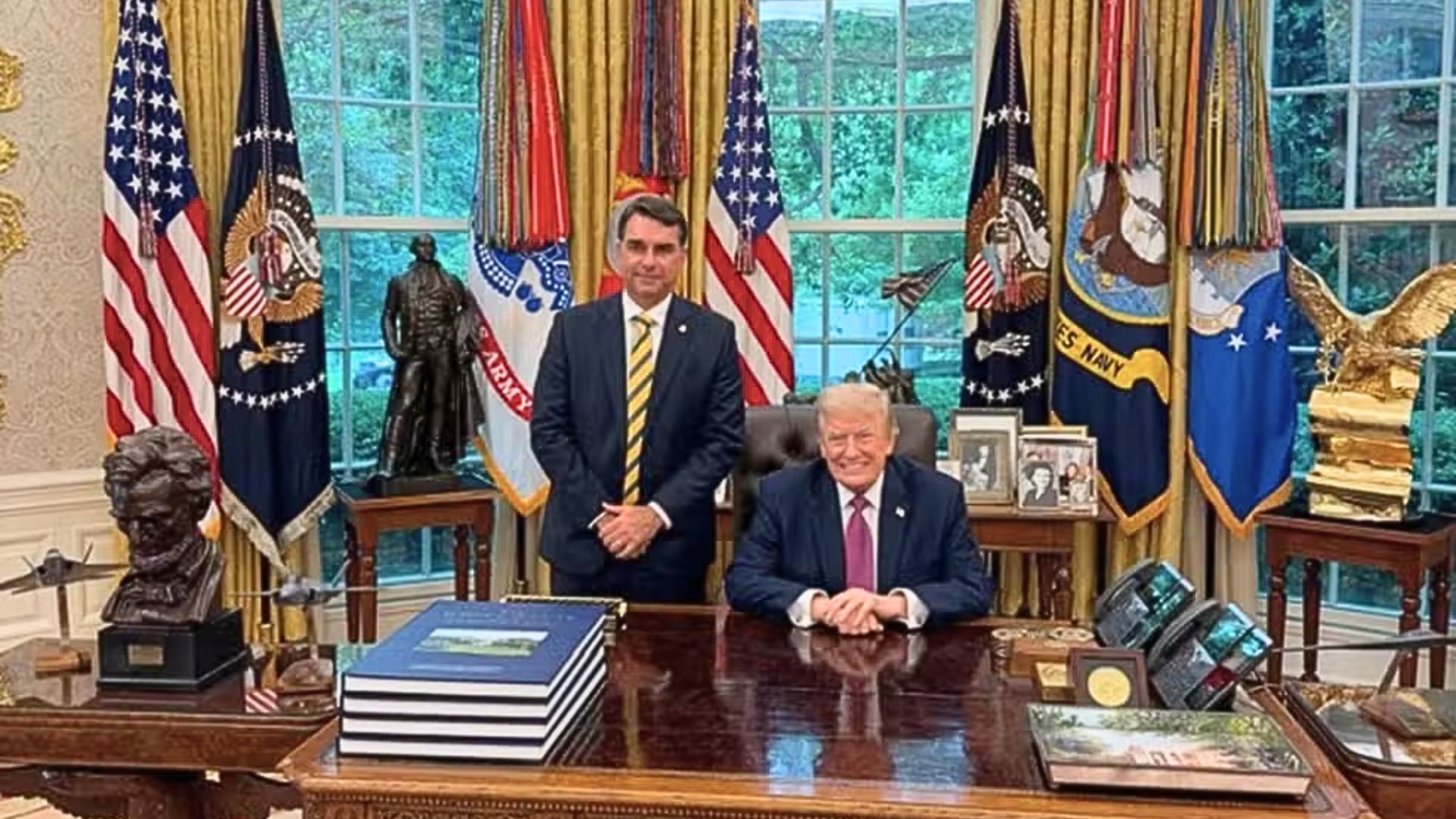Em salão da Casa Branca com bandeira dos EUA, sorriem Donald Trump e Flávio Bolsonaro. Donald está sentado, Flávio em pé ao seu lado