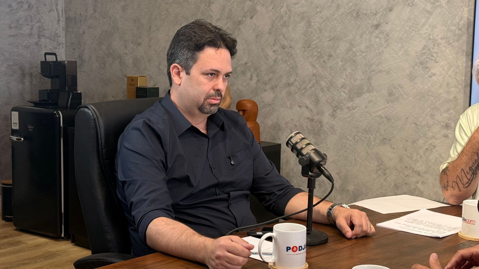 Felipe Alecrim olha para o lado enquanto está sentado em frente a um microfone e uma caneca do PodJá