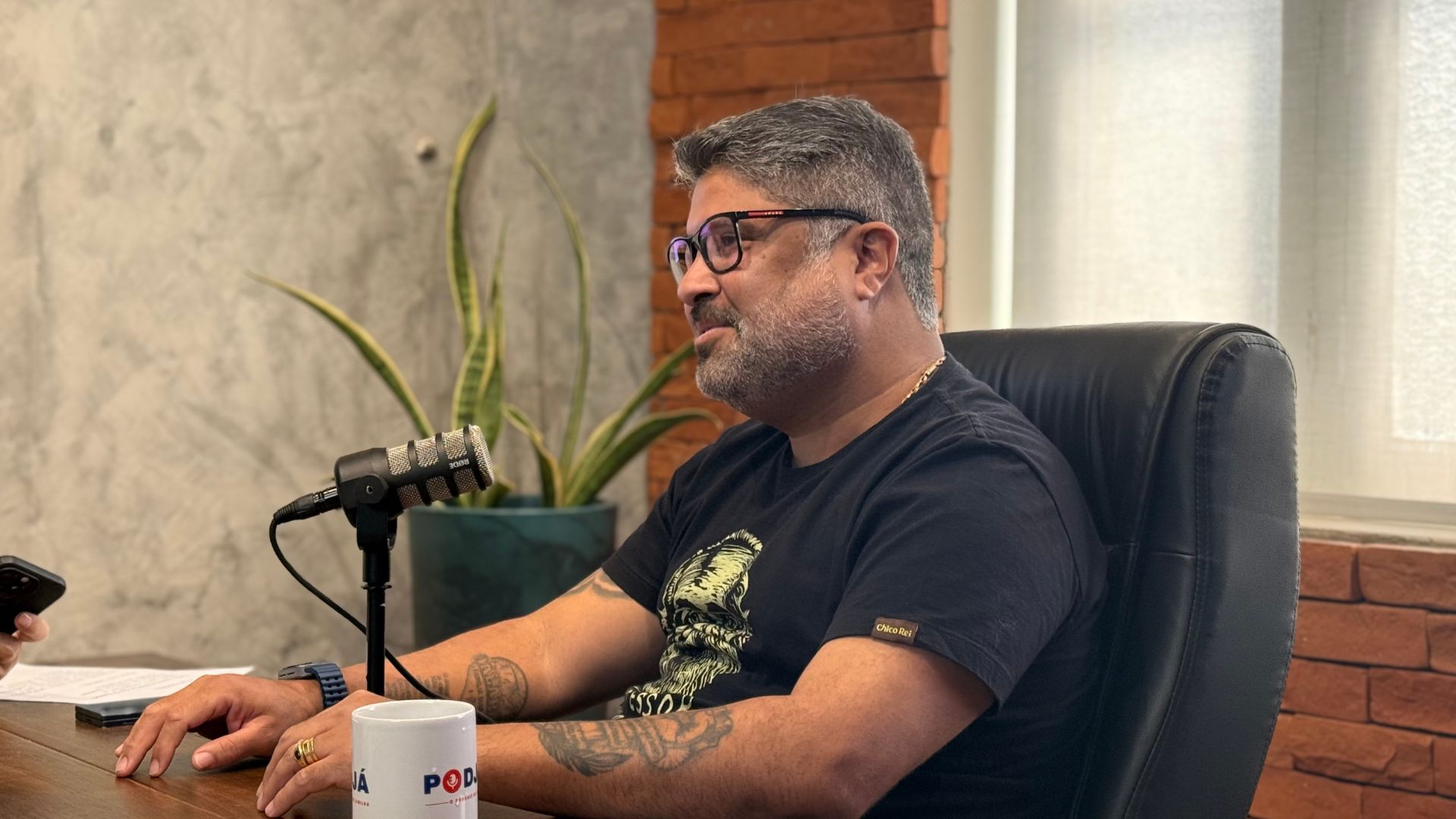 Rinaldo Júnior sorri levemente enquanto olha para o lado, está em frente a um microfone e com caneca do PodJá