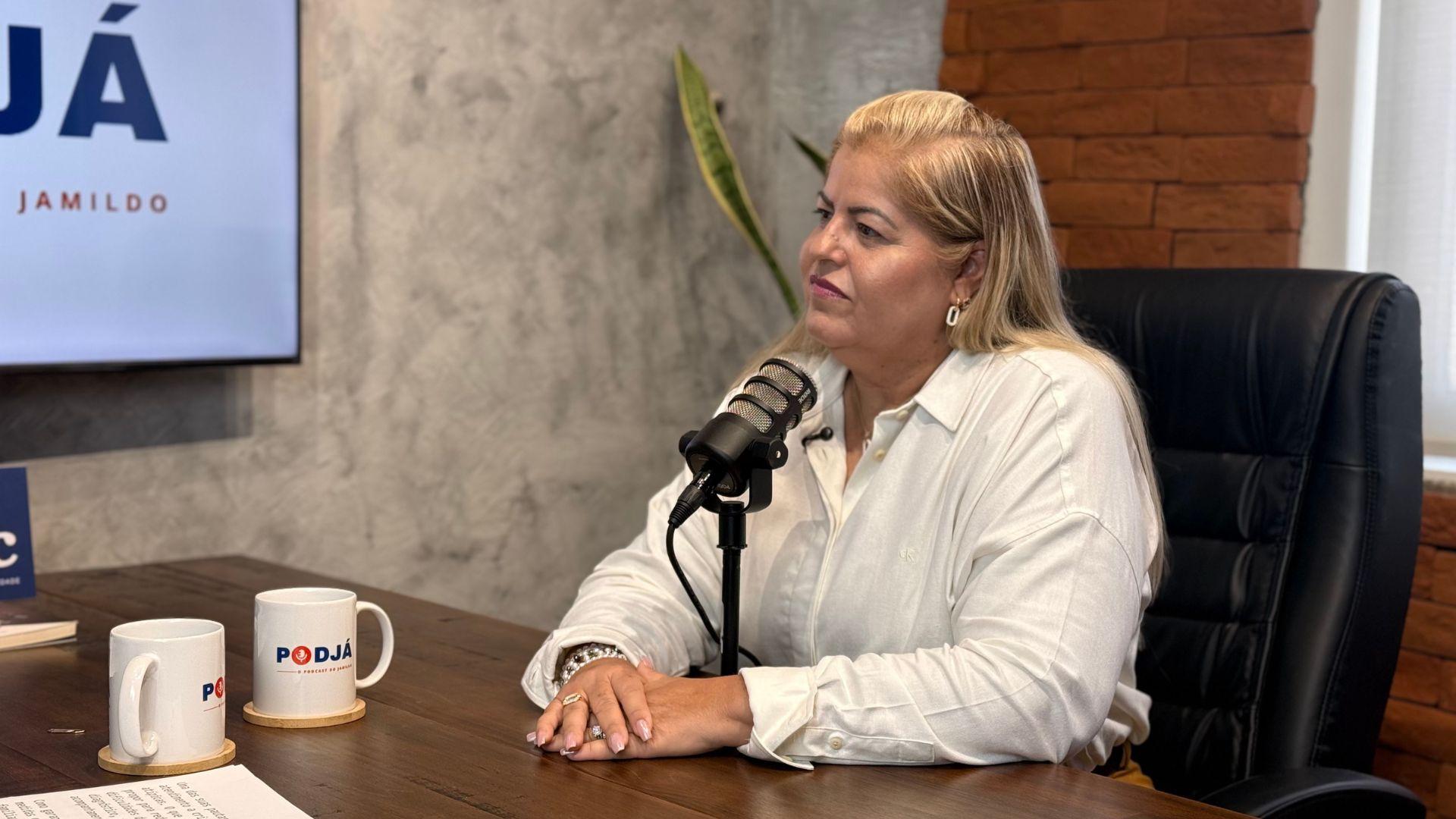 Andrea Medeiros fala sentada em frente a microfone de podcast