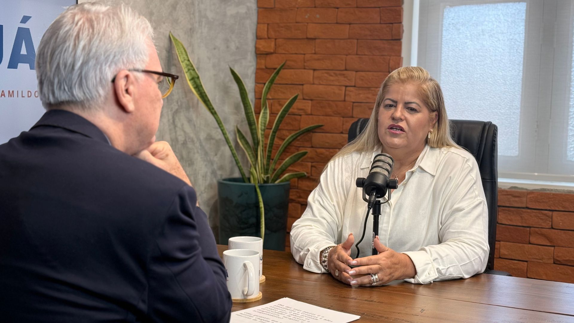 Andrea Medeiros fala sentada em frente a microfone de podcast. Jamildo de costas sentado em frente a Andrea olha para ela