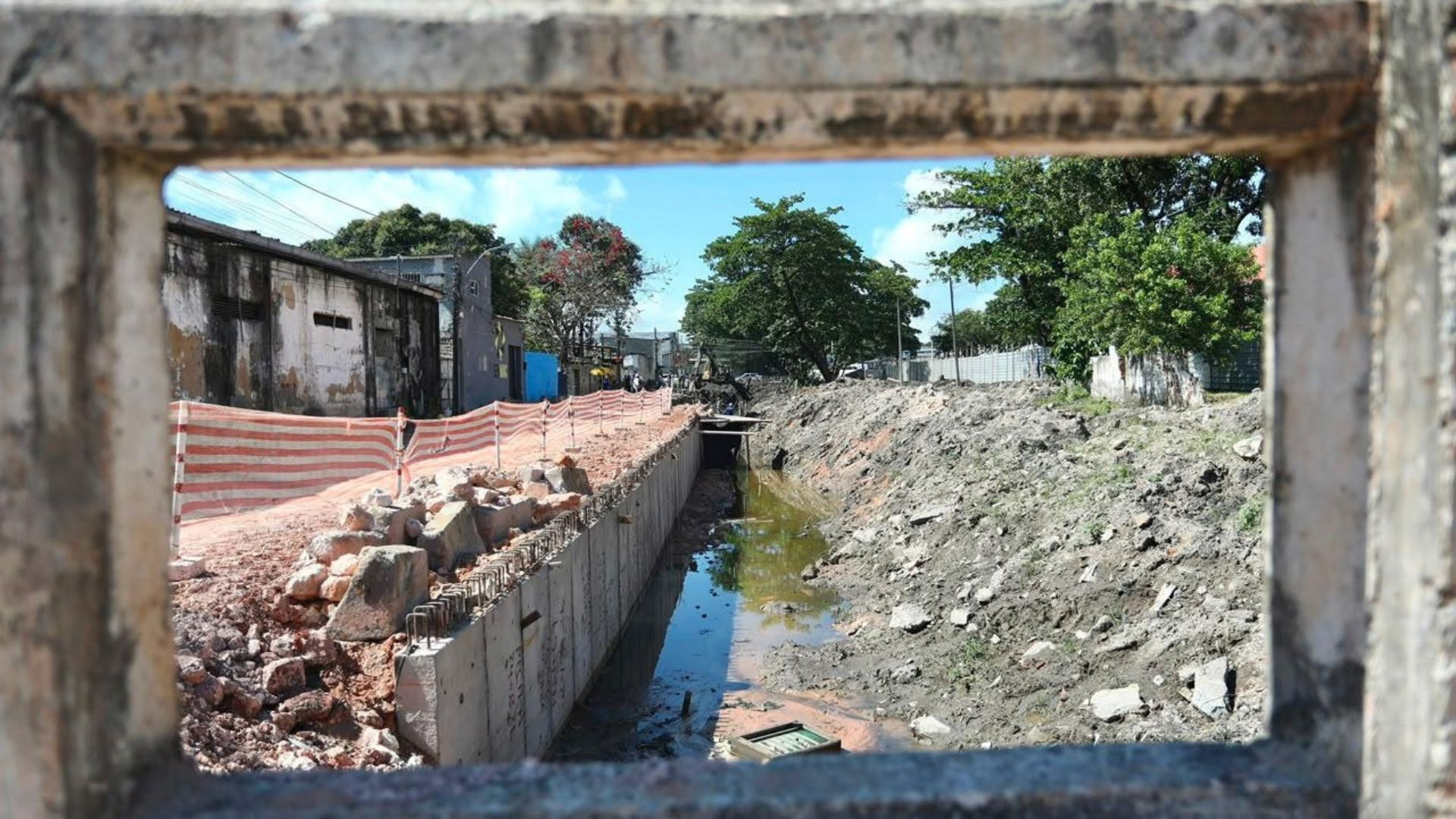 Vista de canteiro de obras com canal em estruturação e contenção de concreto, capturada através de abertura em muro. Ao lado, via de terra com isolamento, vegetação e edificações sob o céu claro.