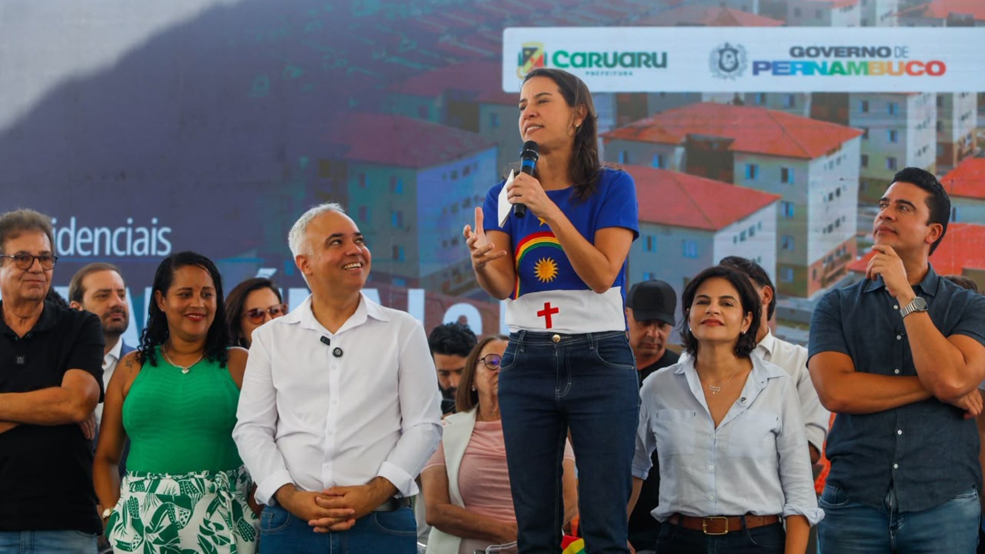 Raquel Lyra com microfone discursando entre o ministro das Cidades, Priscila Krause e lideranças políticas