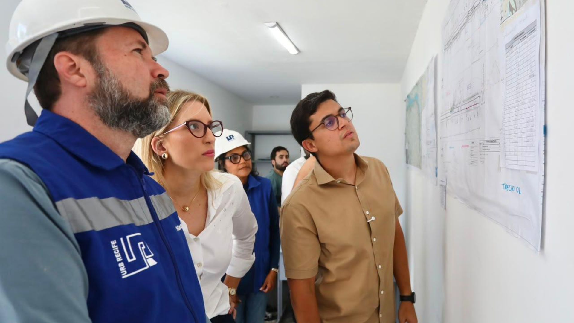 O prefeito Victor Marques, Beatriz Menezes e equipe da Prefeitura do Recife, vestindo crachás e capacete de segurança, analisam juntos mapas e cronogramas de engenharia fixados na parede de escritório de planejamento.